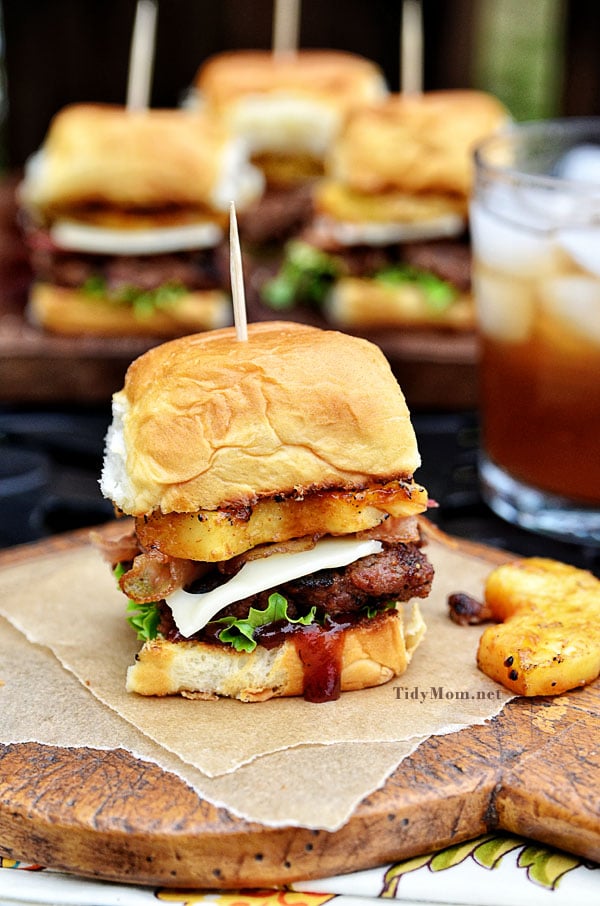 Fire up the grill for this Hawaiian burger recipe. Aloha BBQ Sliders are flavored with BBQ sauce, served on sweet rolls with cheese, pineapple and bacon. Print recipe at TidyMom.net