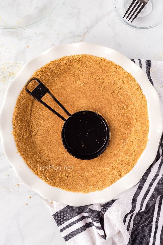 graham cracker crust being pressed into dish with measuring cup