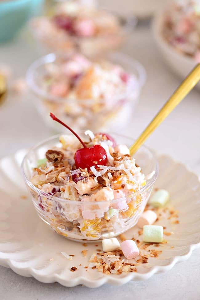 serving of Ambrosia Salad in a clear cup with a cherry on top and a gold spoon