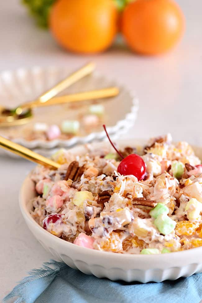 White bowl full of ambrosia salad with a cherry on top and blue napkin