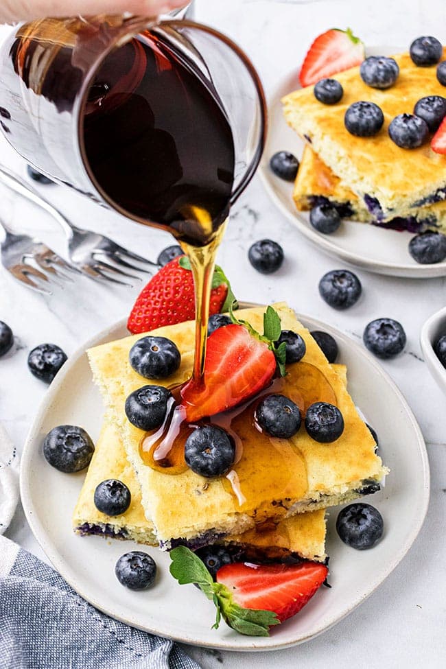 a stack of oven baked pan sheet pancakes on a plate with fresh berries and syrup being poured over them.
