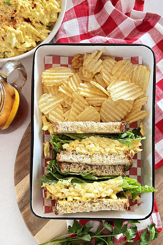 Classic Egg Salad Sandwich in a white tray with potato chips and red checked napkin