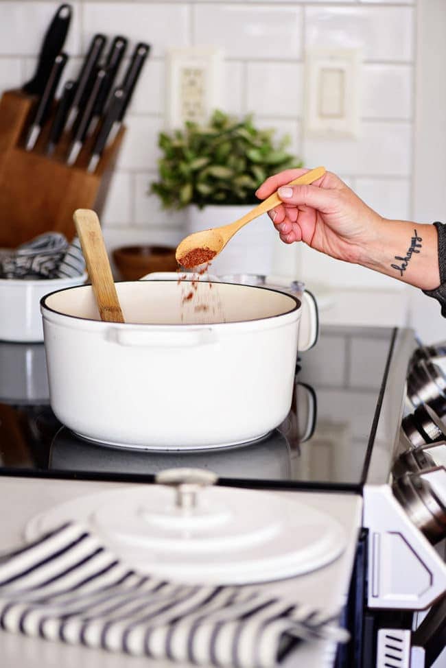 white stock pot on a stove with woman sprinkling chili seasoning with a wooden spoon