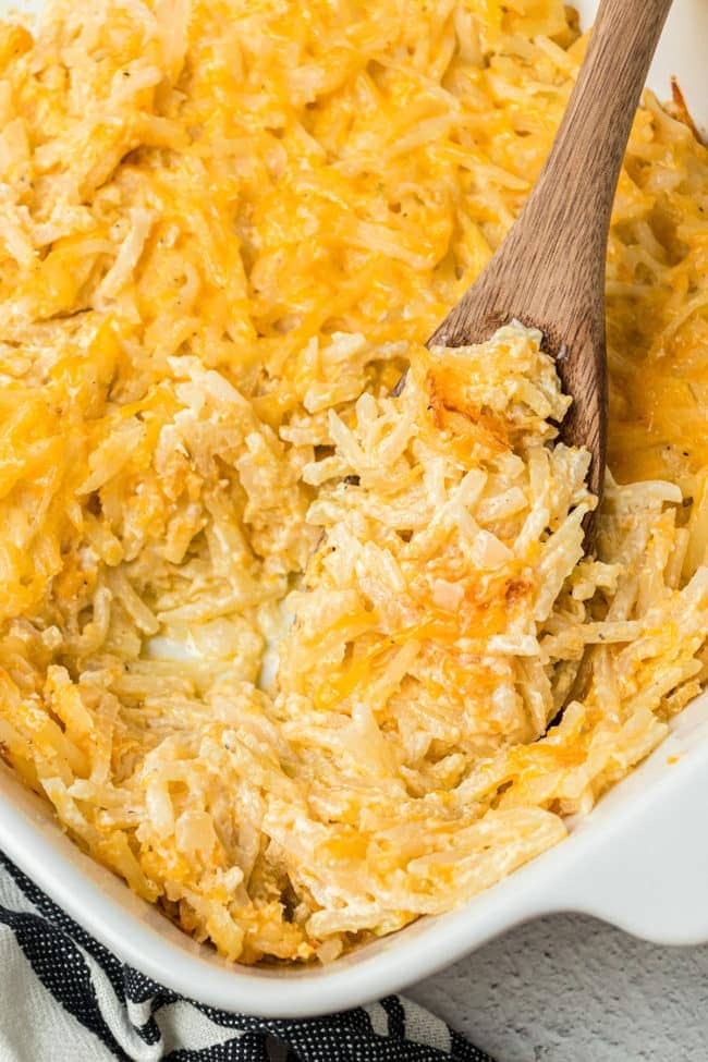 close up of cheesy hashbrown casserole in a baking dish with a wooden spoon