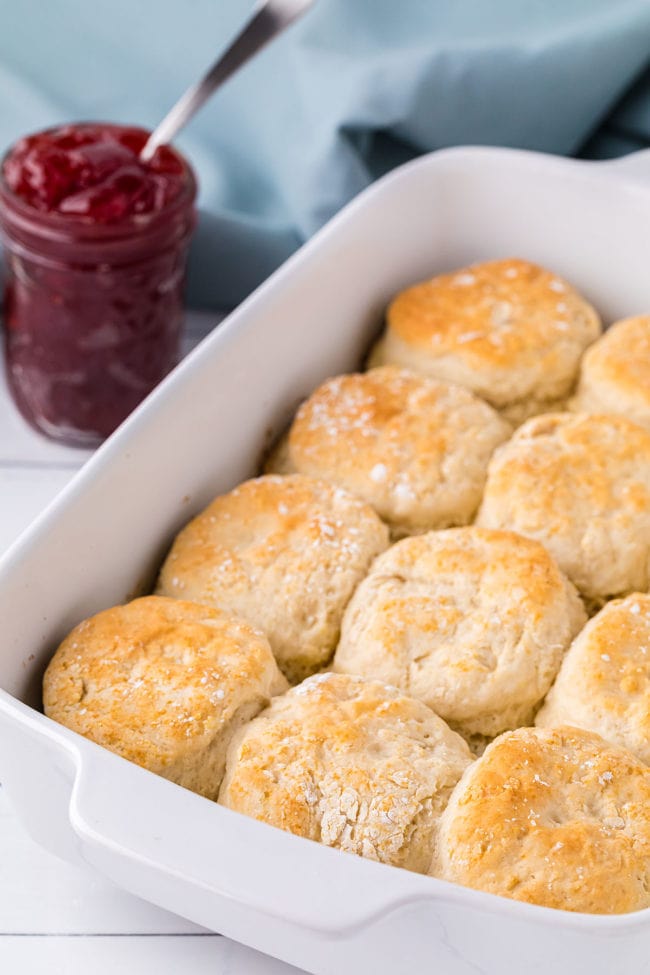 homemade biscuits in a white dish with a jar of jelly and blue towel in backgroung