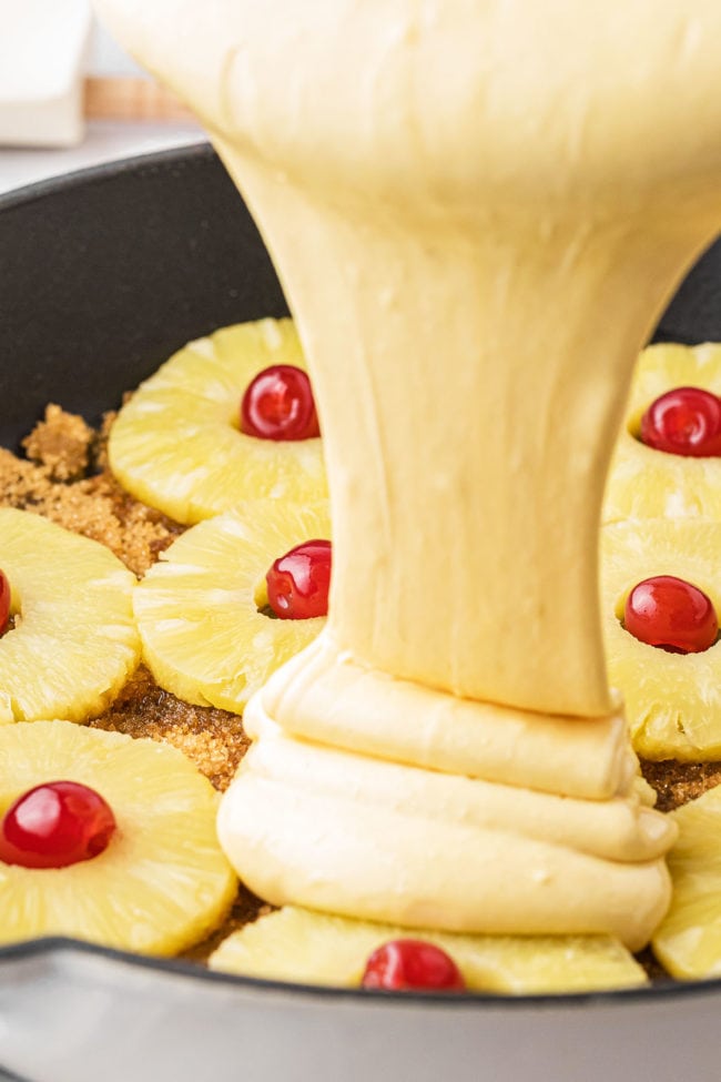 pouring cake batter over pineapple and cherries
