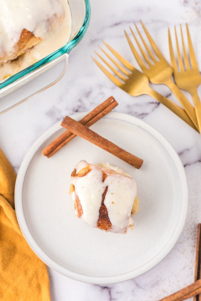 homemade cinnamon roll on a plate with 2 cinnamon sticks and gold forks on the table next to the plate.