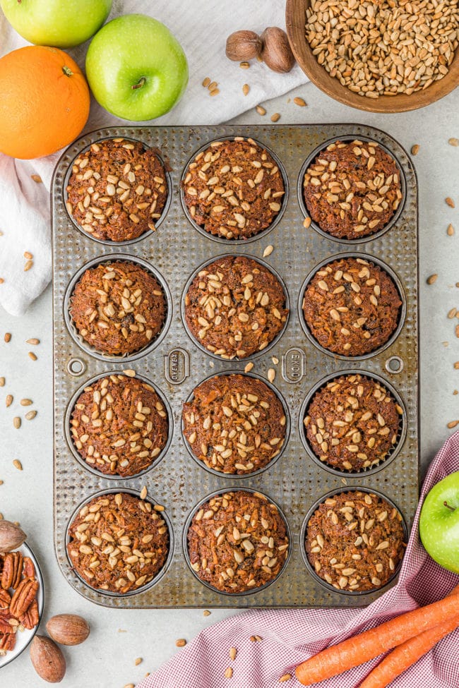 muffins in a pan, with carrots, apples, orange and nuts on the counter around the pan