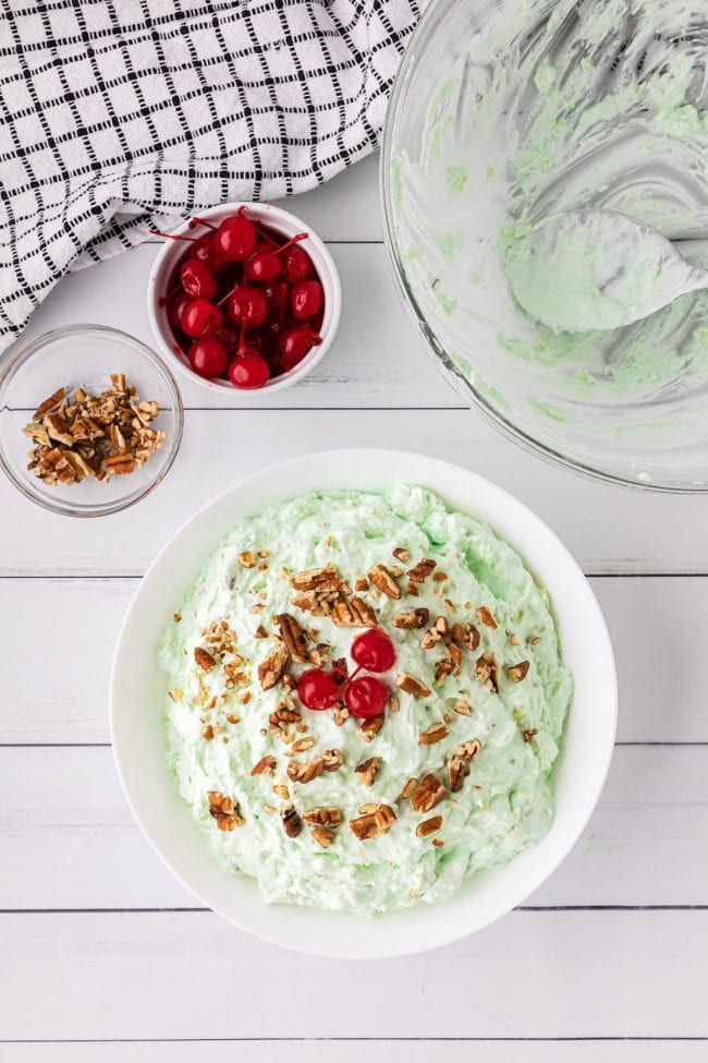 a white bowl of green fluff salad and little bowls of chopped pecans and cherries next to the bowl.