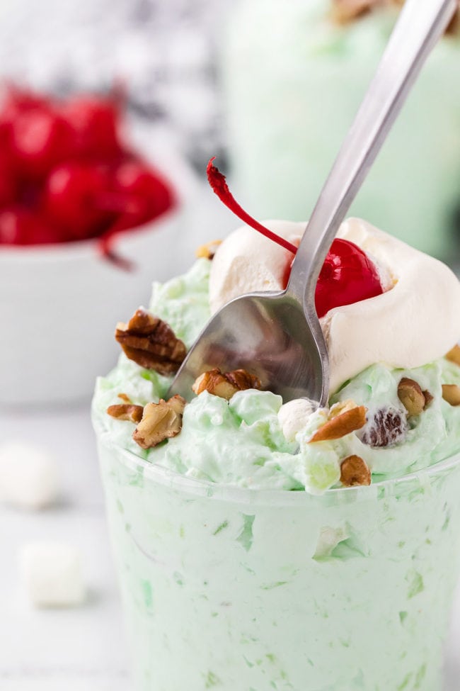 a parfait cup of pistachio salad with a spoon digging in and a bowl of cherries in the background.