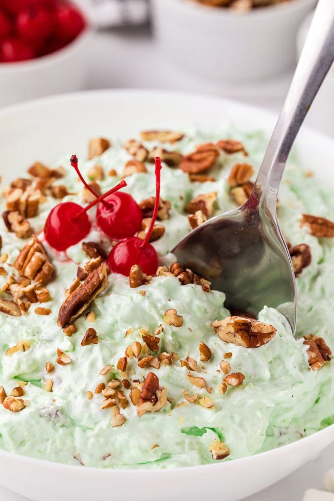 green pistachio fluff salad  in a white bowl with a spoon and chopped nuts and cherries on top