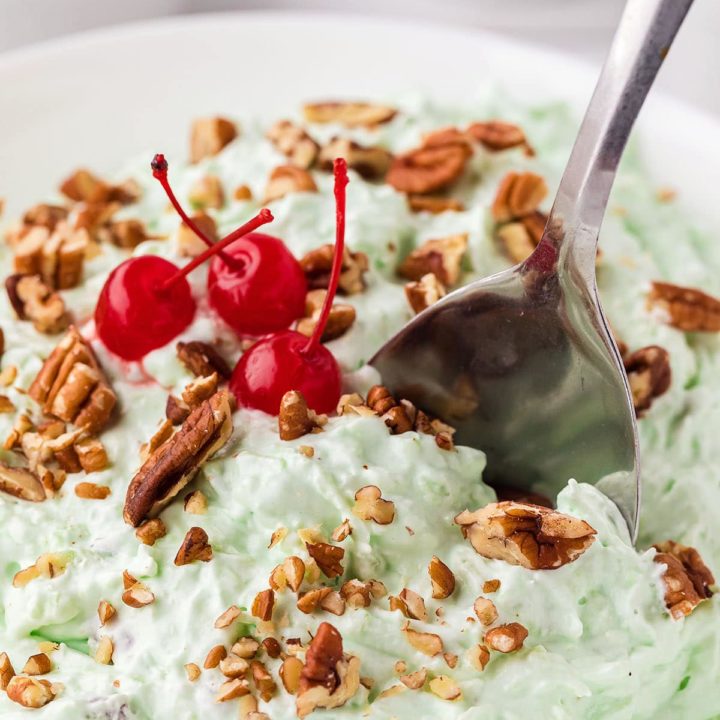 green pistachio fluff salad in a white bowl with a spoon and chopped nuts and cherries on top