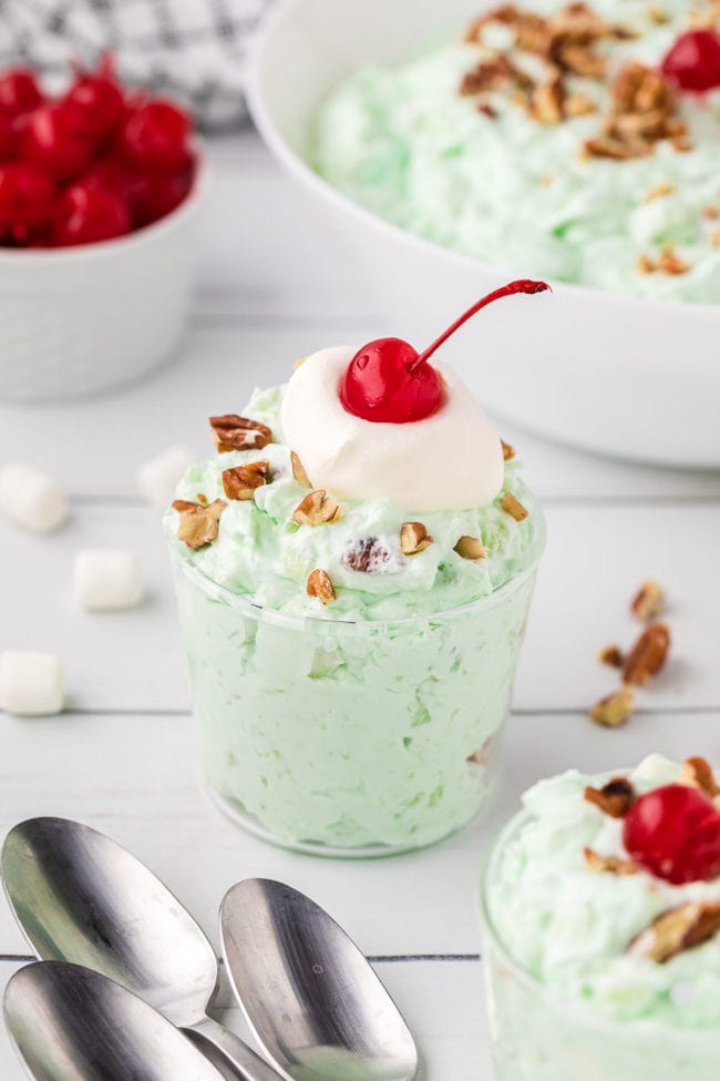 pistachio salad in a dessert cup with spoons on the table and cherries and a bowl of green fluff in the background