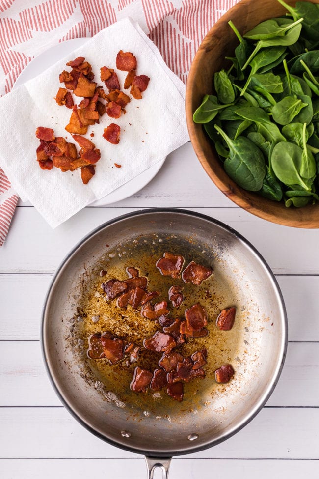 frying bacon in a small skillet with a bowl of baby spinach and a paper towel with cooked bacon
