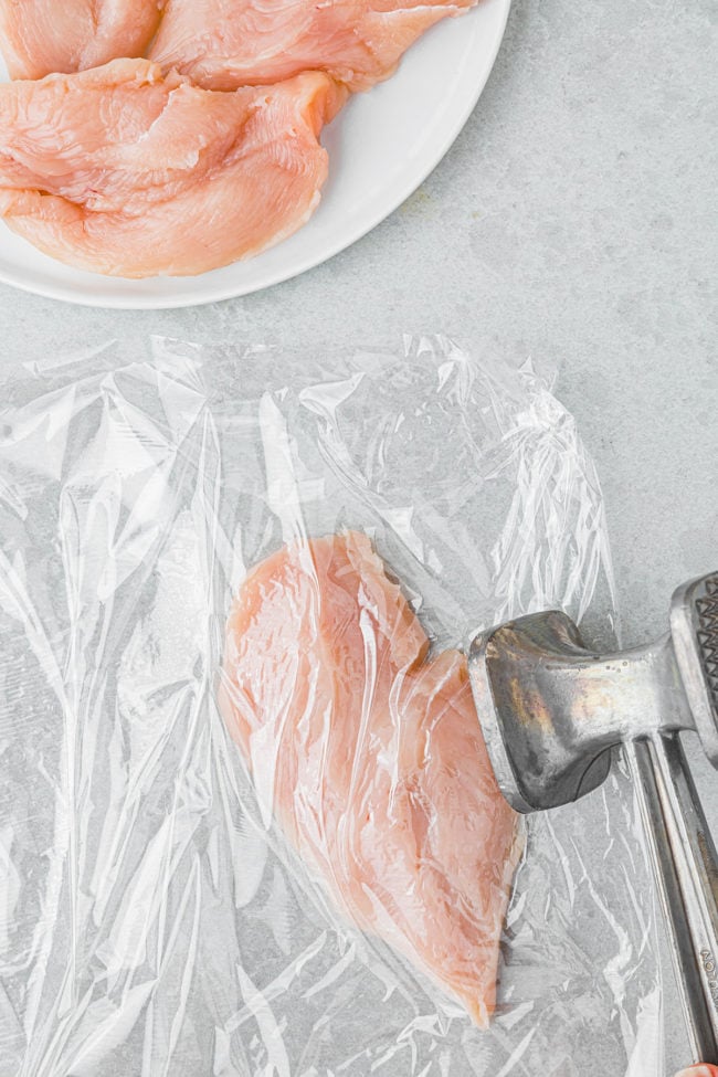 pounding a chicken breast covered in plastic wrap on a counter