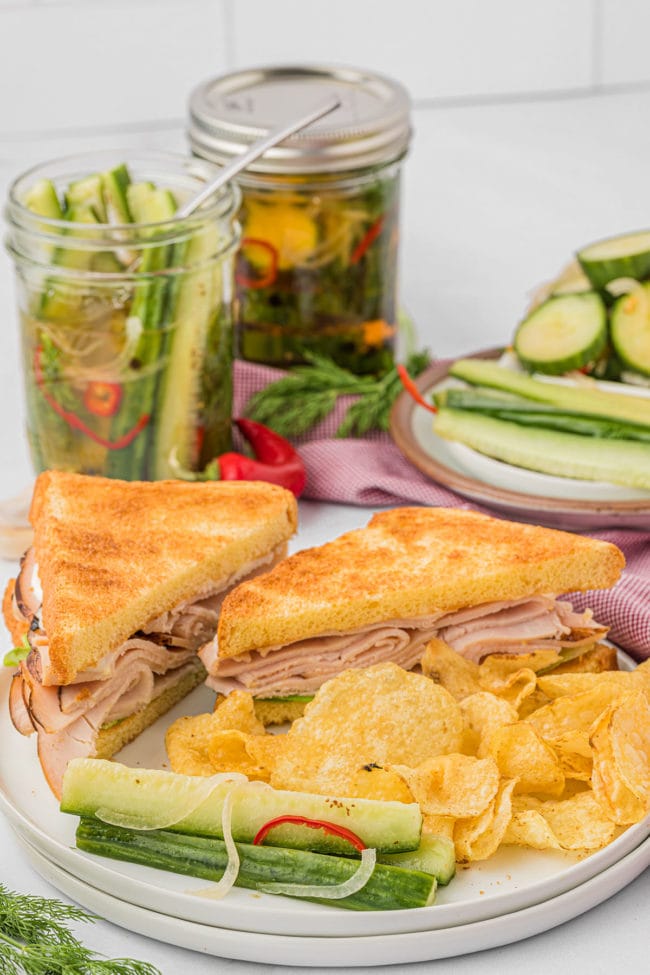 a sandwich on a plate with pickles and chips and jars of homemade quick pickles in the background