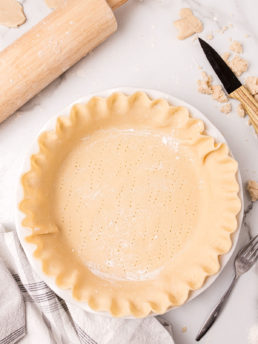 unbaked pie crust with crimped edges in a white pie plate next to a wood rolling pin