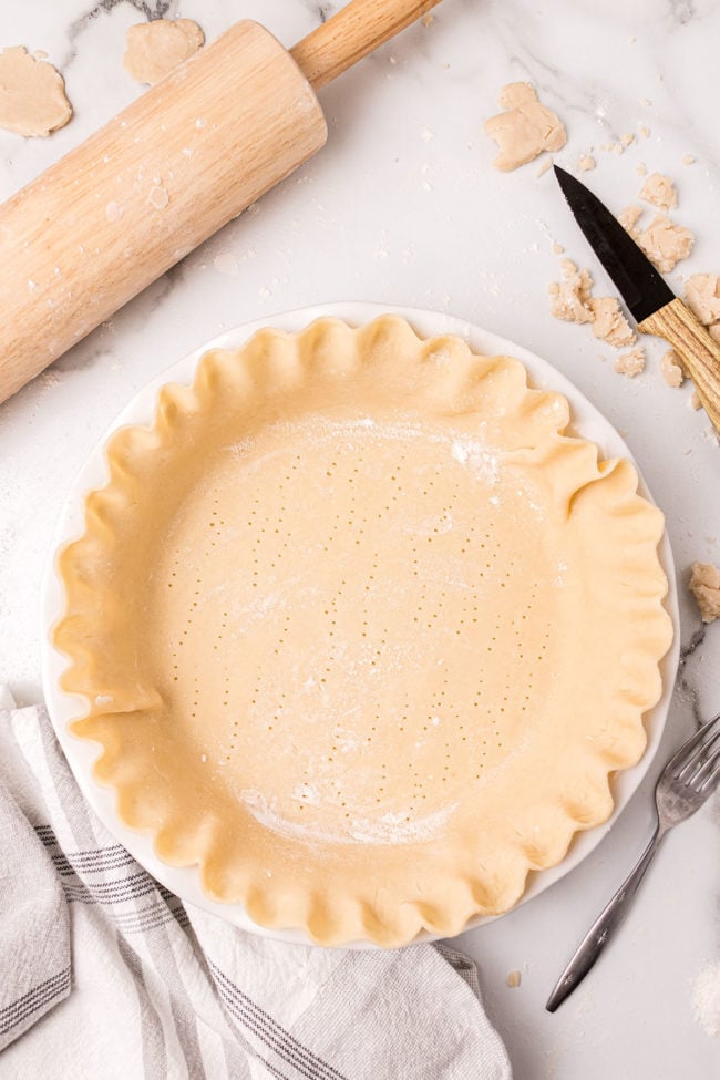 unbaked pie crust with crimped edges in a white pie plate next to a wood rolling pin