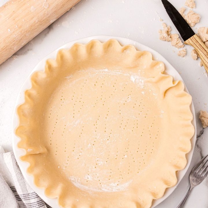 unbaked pie crust with crimped edges in a white pie plate next to a wood rolling pin