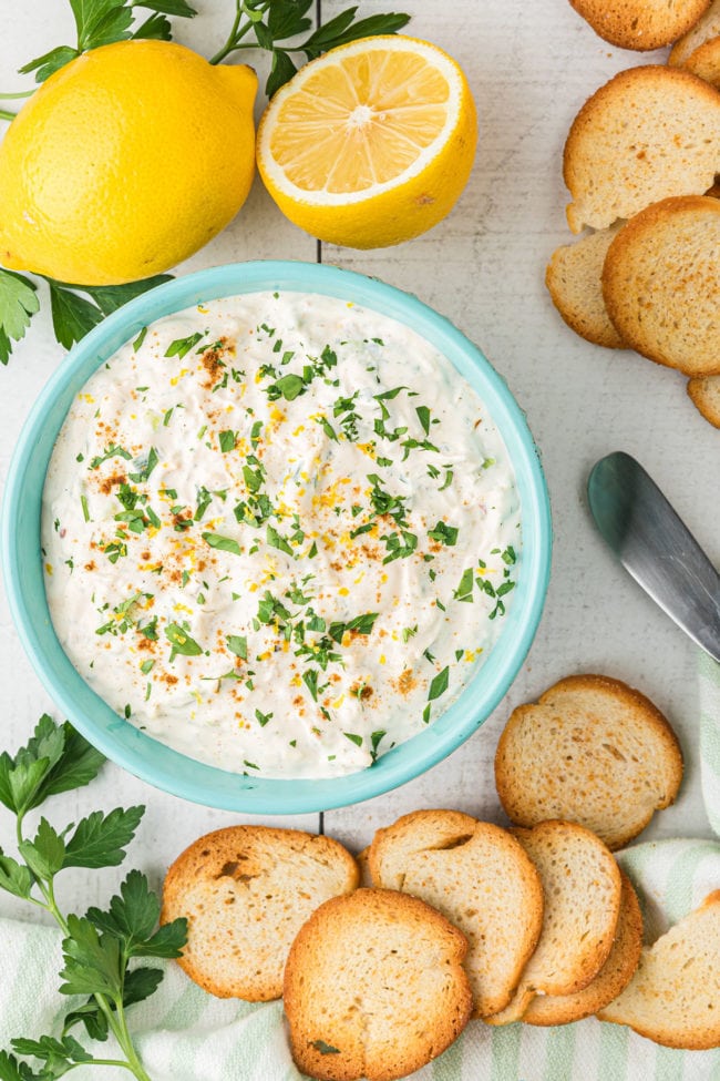 overhead shot of crab dip in a blue bowl surrounded by baguette crackers, fresh lemon and parsley