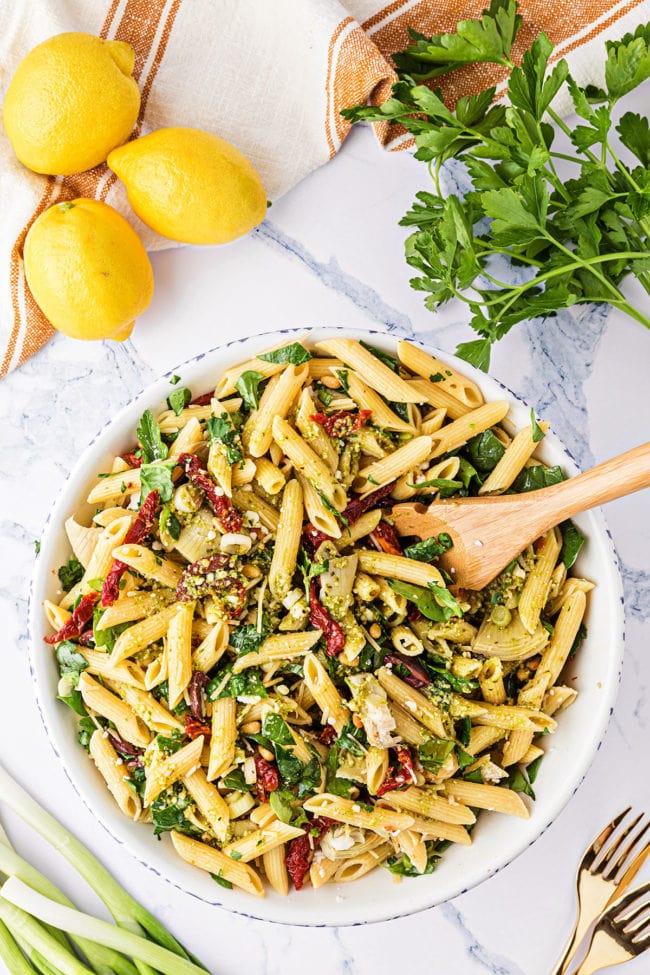 Bowl of Greek Pasta Salad with a wooden spoon. Fresh parsley and lemons in the background