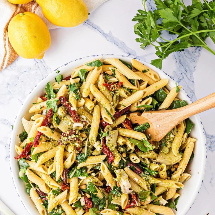 Bowl of Greek Pasta Salad with a wooden spoon. Fresh parsley and lemons in the background