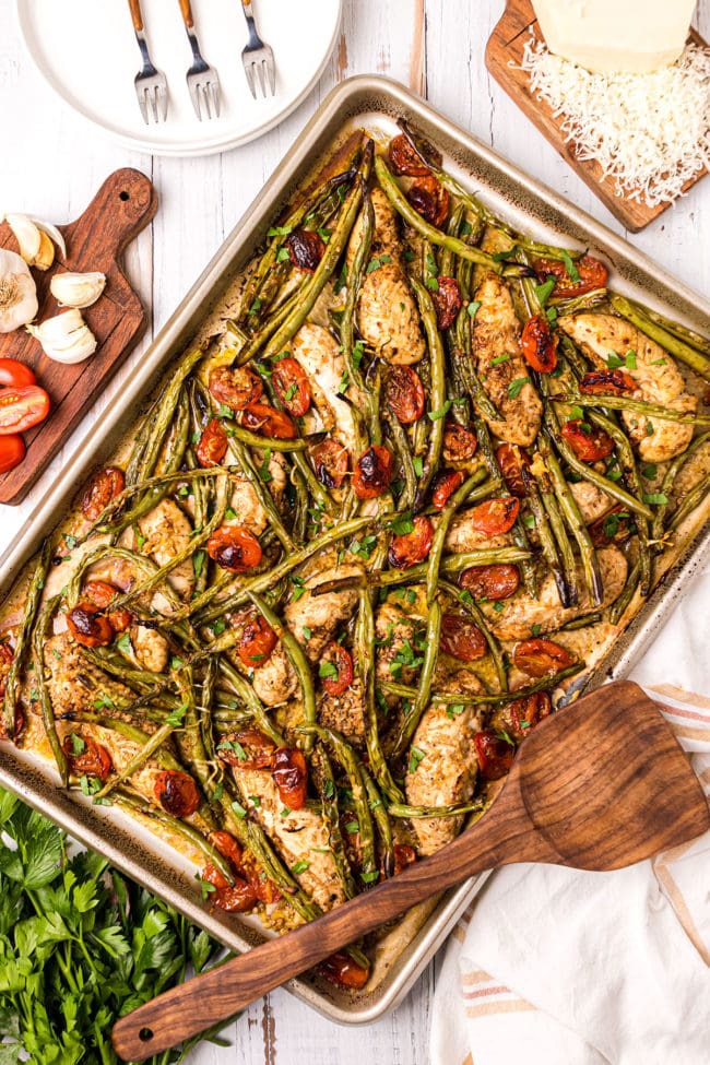 chicken tenders, green beans and tomatoes on a sheet pan with a wooden spatula