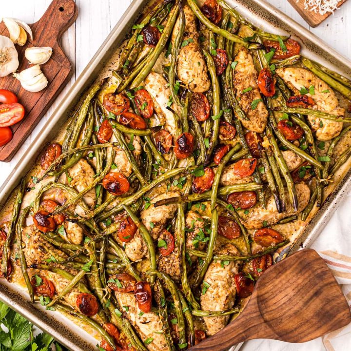 chicken tenders, green beans and tomatoes on a sheet pan with a wooden spatula