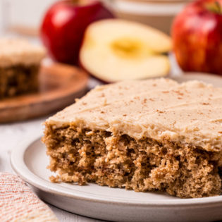 slice of cake on a plate. Red apples in the background.