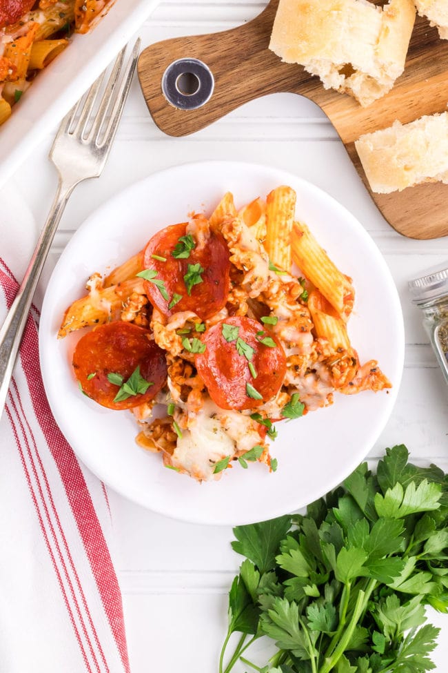 a dish with pasta and pepperoni in a red sauce on a white plate with fresh parsley next to it