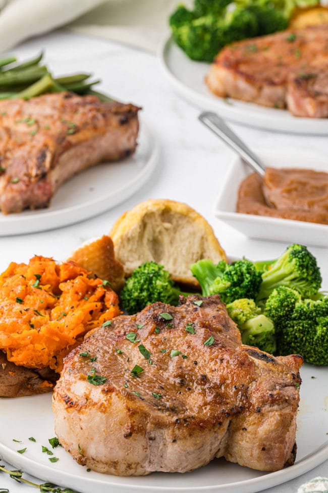 apple butter glazed pork chops on a plate with a baked sweet potato, broccoli and a dinner roll