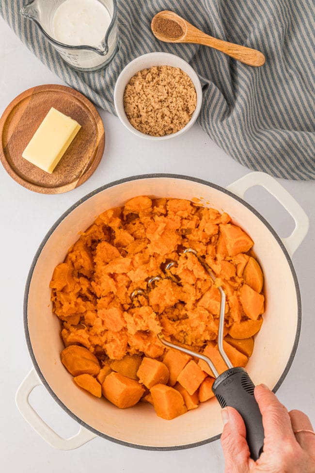 mashing cooked sweet potatoes with a hand potato masher in a white dutch oven