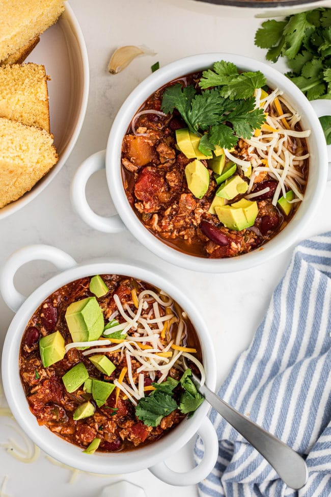 2 bowls of turkey chili topped with avocado, cheese and fresh herbs