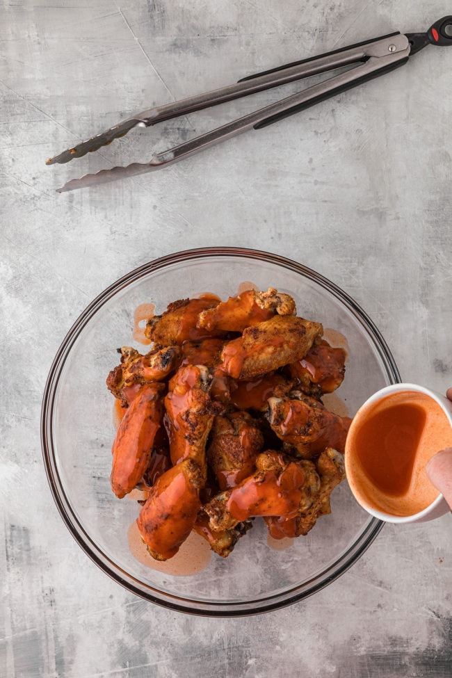buffalo sauce on baked chicken wings in a glass mixing bowl.