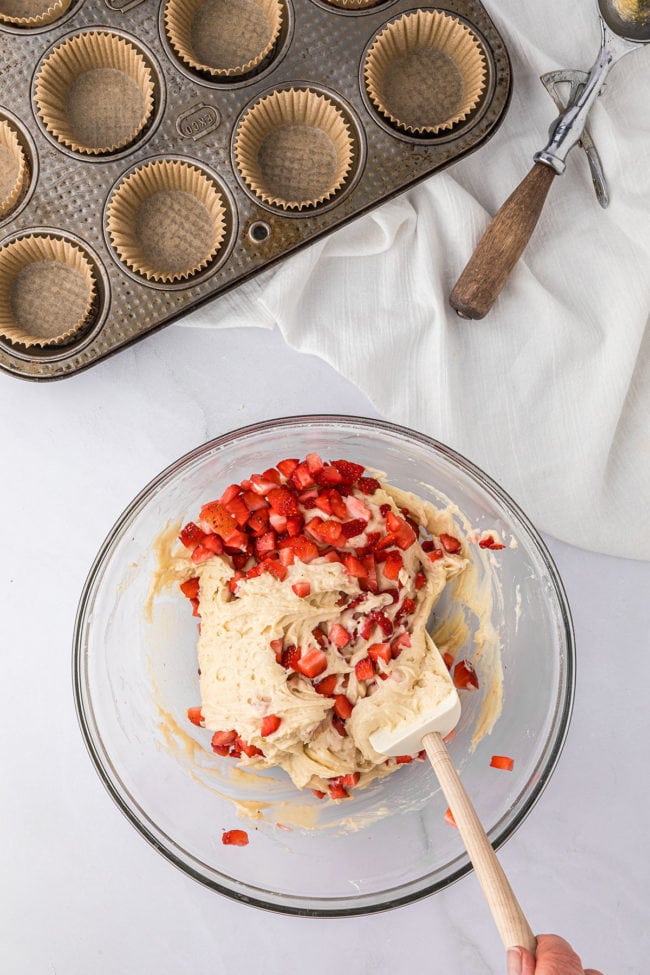 fresh strawberries in a muffin batter in a glass mixing bowl with a rubber spatula