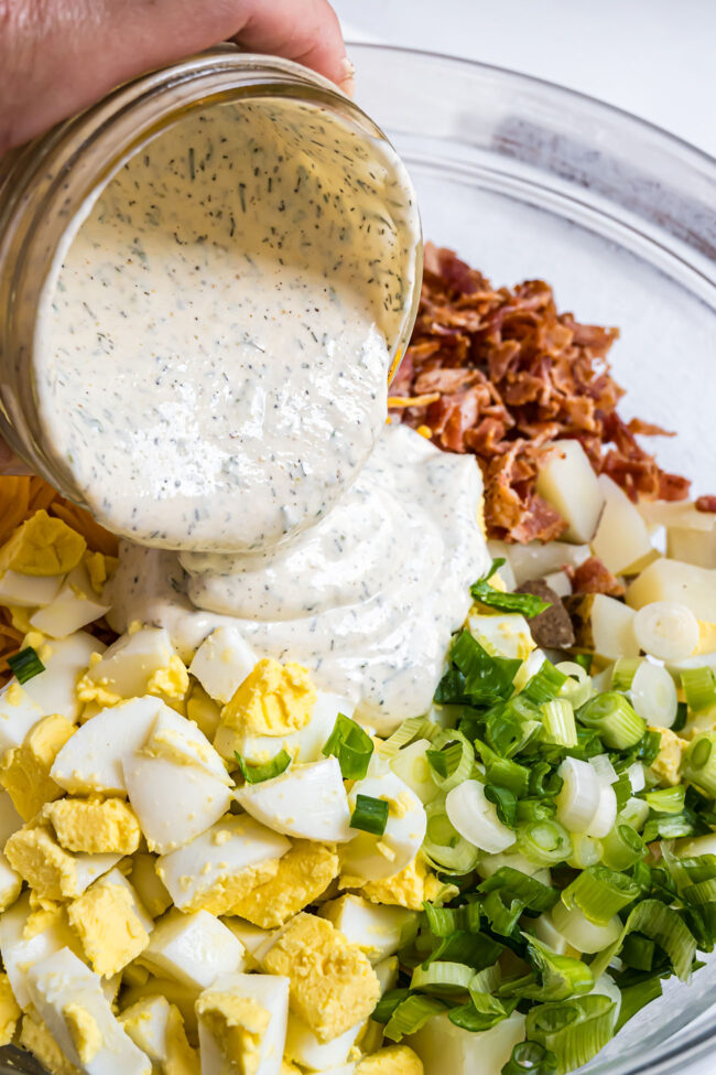 pouring ranch dressing over a bowl of Loaded Baked Potato Salad ingredients