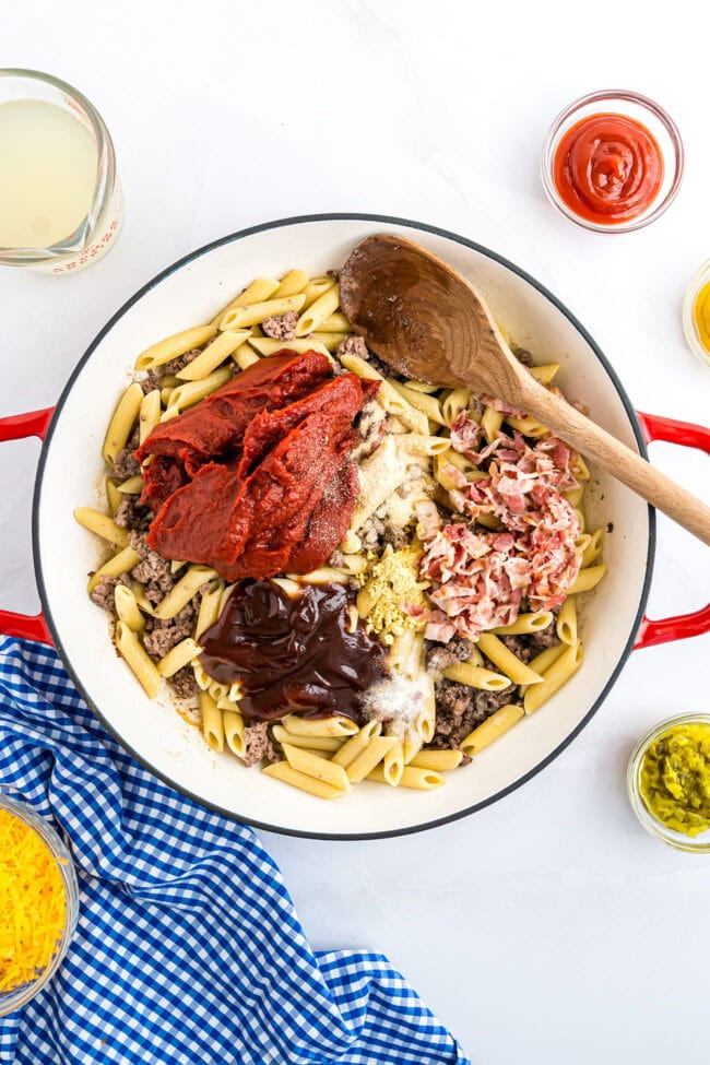 Skillet with cooked ground beef, penne pasta, diced bacon, tomato paste, and BBQ sauce ready to be stirred together.