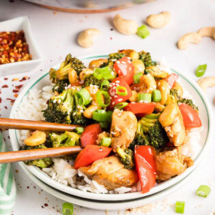 Cashew Chicken Stir Fry plated and served over white rice with chopsticks — tender chicken, crisp vegetables, and cashews coated in a glossy, savory-sweet sauce.