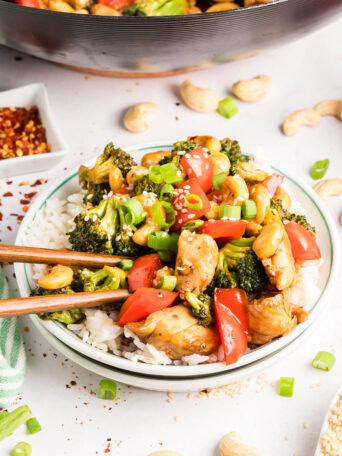 Cashew Chicken Stir Fry plated and served over white rice with chopsticks — tender chicken, crisp vegetables, and cashews coated in a glossy, savory-sweet sauce.