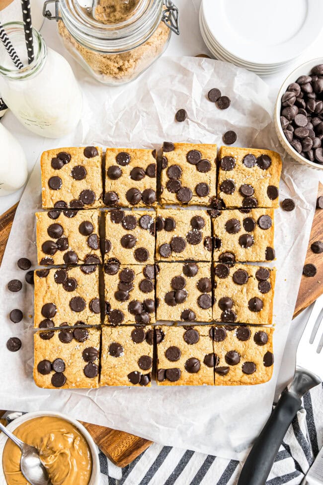 Overhead shot of cut peanut butter blondies arranged in a square on parchment paper, surrounded by milk and baking ingredients.