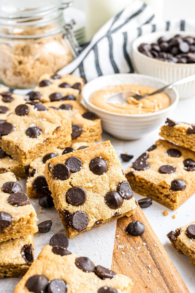 Cut squares of peanut butter blondies on a tabletop with a cutting board and bowls of peanut butter, chocolate chips, and brown sugar in the background.