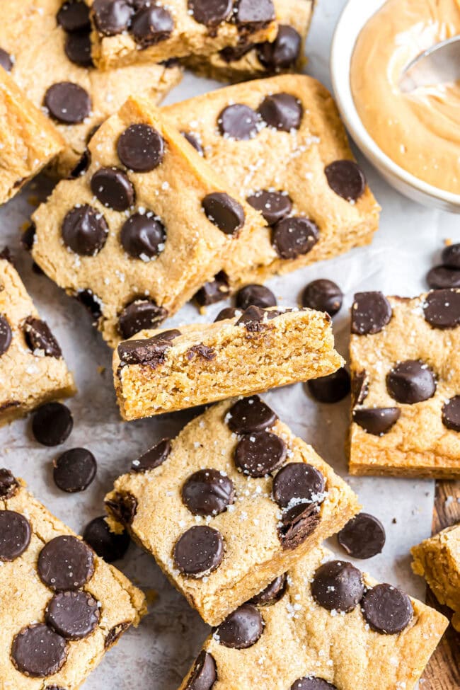 Close-up flatlay of peanut butter blondie bars with a bowl of peanut butter and one bar turned on its side showing the soft, chewy center.