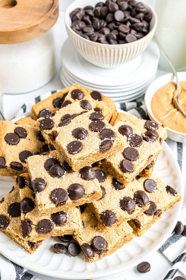 White plate stacked with peanut butter blondies in a pyramid, with chocolate chips and peanut butter bowls blurred in the background.
