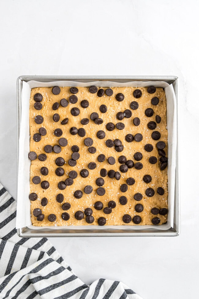 Blondie batter spread in a parchment-lined pan with chocolate chips and flaky sea salt sprinkled on top before baking.