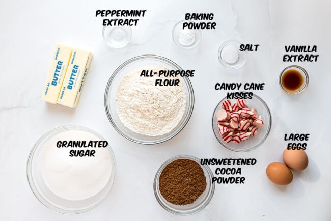 Ingredients for Chocolate Peppermint Kiss Cookies laid out on a counter, including cocoa powder, flour, sugar, butter, eggs, and candy cane Kisses.