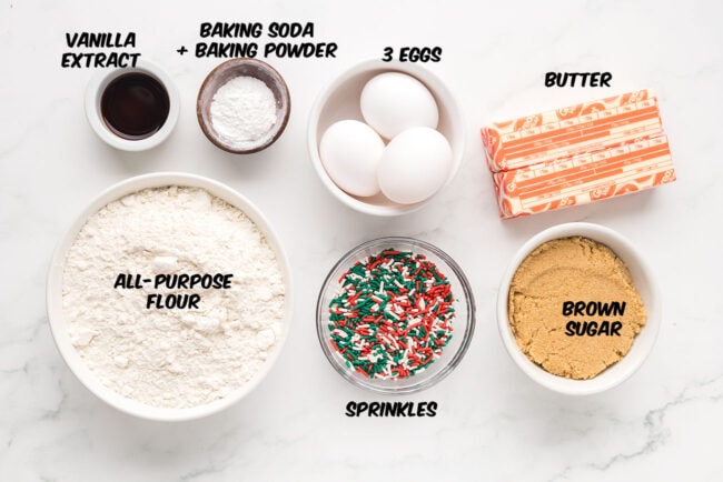 Ingredients laid out on a counter to make Christmas Blondies, including butter, brown sugar, eggs, flour, and holiday sprinkles.