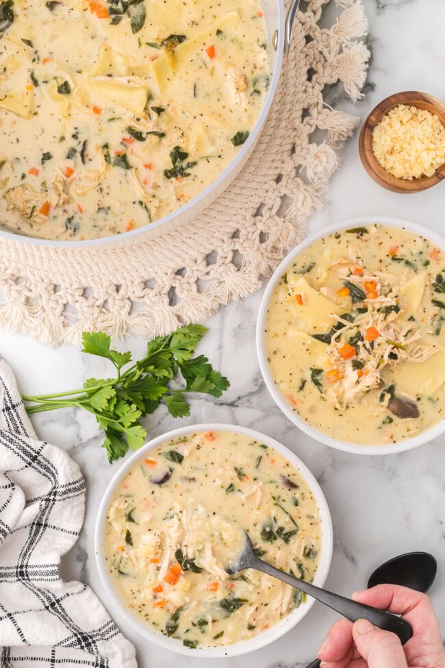 Bowls of creamy white lasagna soup with chicken and spinach, a Dutch oven of soup nearby, and a hand holding a spoon over one bowl.