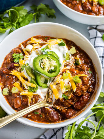 Close-up bowl of Easy Cowboy Chili topped with sour cream, shredded cheddar cheese, sliced jalapeños, and green onions.