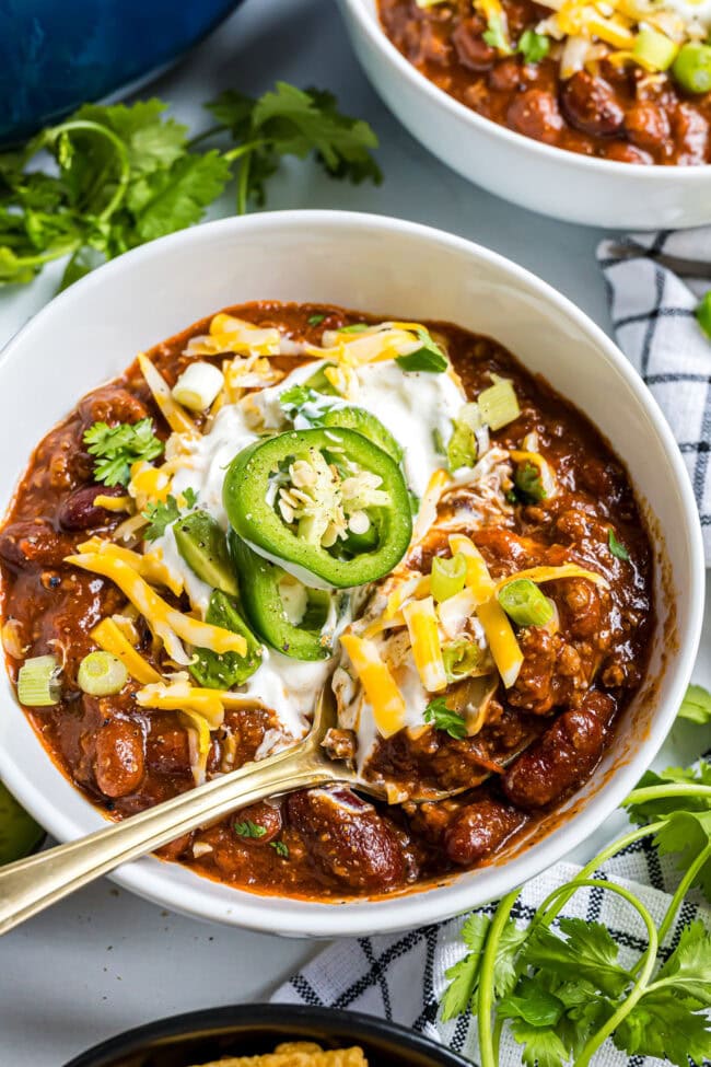Close-up bowl of Easy Cowboy Chili topped with sour cream, shredded cheddar cheese, sliced jalapeños, and green onions.
