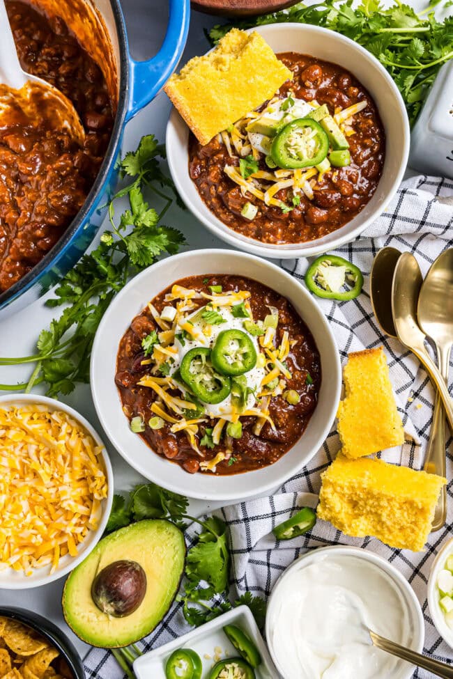 Overhead view of two bowls of Easy Cowboy Chili topped with shredded cheddar cheese, sour cream, sliced jalapeños, and green onions, served with cornbread.
